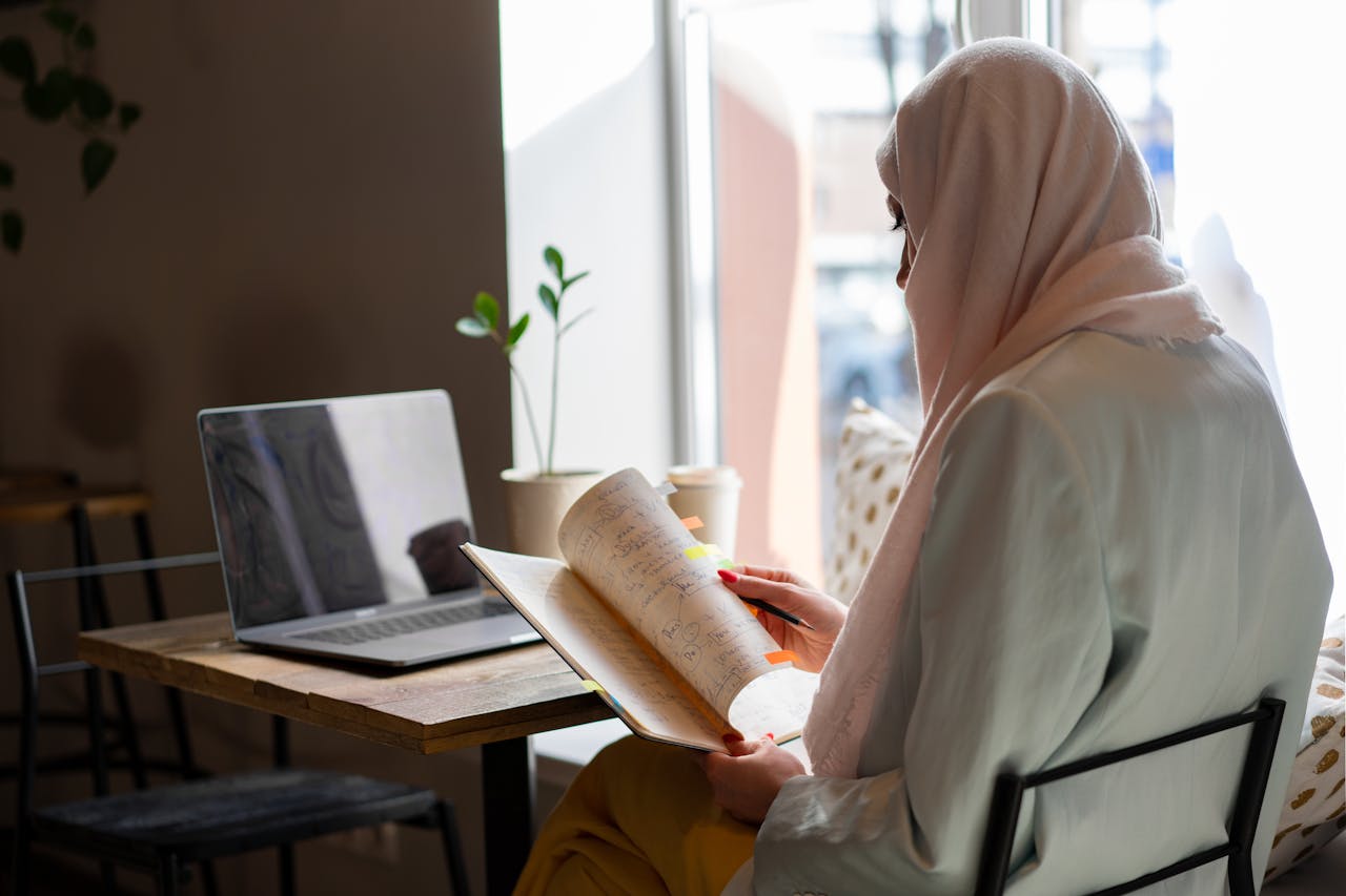 A woman in a hijab sits indoors by a window, reading a book next to a laptop on the table.